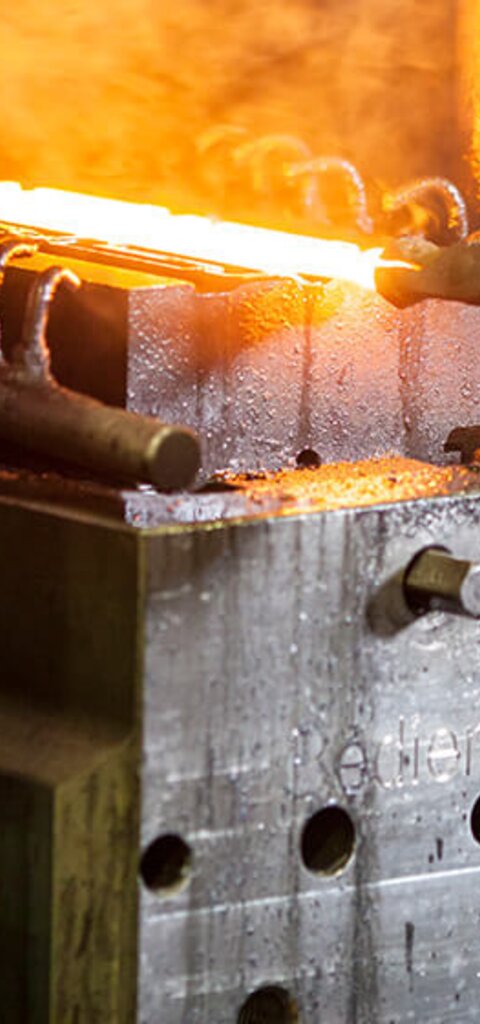 Ein Mann von hinten mit grün-kariertem Hemd hält mit einer Schmiedezange ein glühendes Bauteil, das auf dem Untergesenk der Schmiede liegt - Schmiedetechnik bei RÜBIG