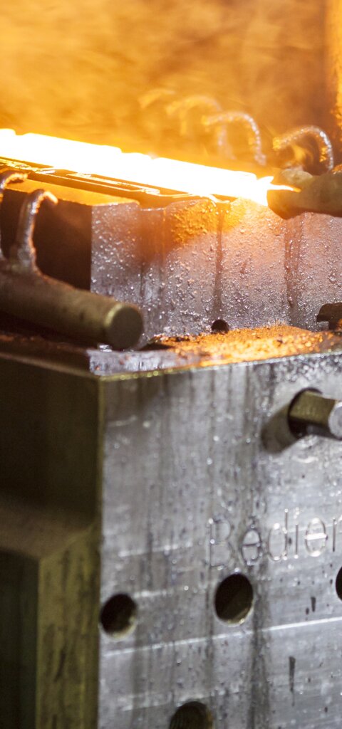 A man in a green checkered shirt holds a glowing forged part with tongs on the lower die – forging technology at RÜBIG