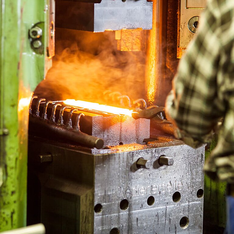 Ein Mann von hinten mit grün-kariertem Hemd hält mit einer Schmiedezange ein glühendes Bauteil, das auf dem Untergesenk der Schmiede liegt - Schmiedetechnik bei RÜBIG