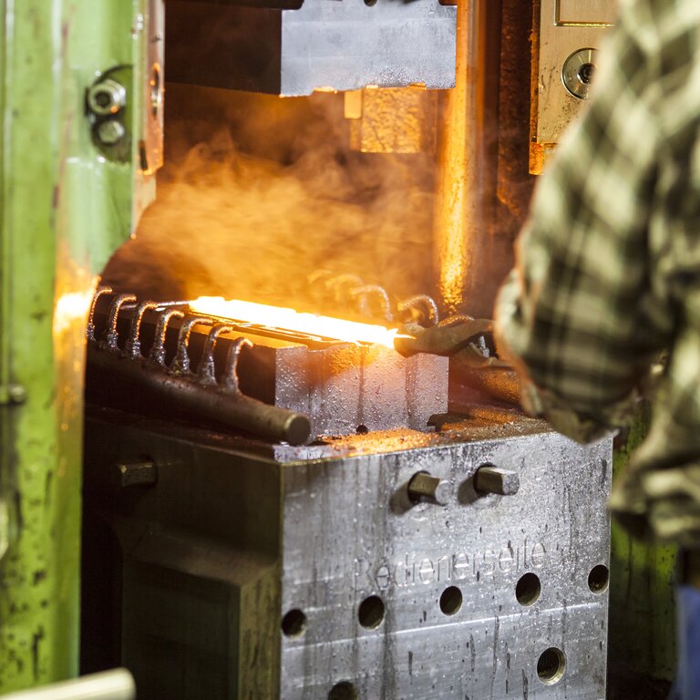 Ein Mann von hinten mit grün-kariertem Hemd hält mit einer Schmiedezange ein glühendes Bauteil, das auf dem Untergesenk der Schmiede liegt - Schmiedetechnik bei RÜBIG