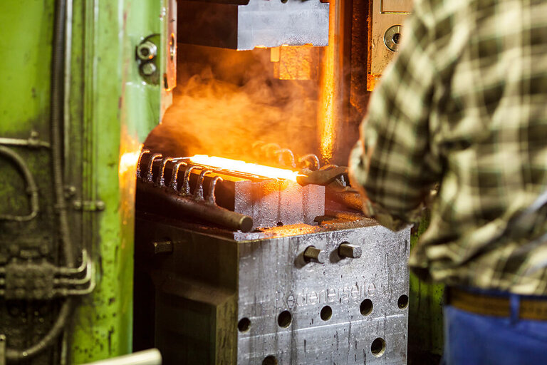 A man in a green checkered shirt holds a glowing forged part with tongs on the lower die – forging technology at RÜBIG