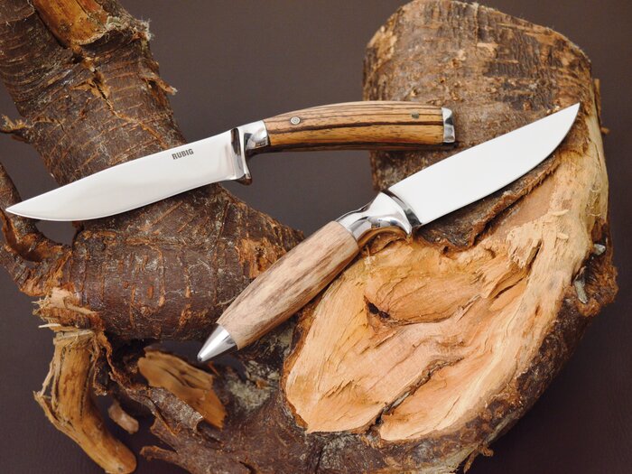 Das RÜBIG-Jagdmesser Genius und das RÜBIG-Jagmesser Helios liegen auf einem Stück Holz. Der Griff besteht jeweils aus Holz.