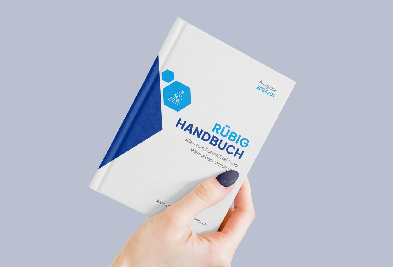 Eine Frauenhand mit blau lackierten Fingernägeln hält ein RÜBIG-Handbuch. Das Buch ist weiß mit blauen, eckigen, geometrischen Farbakzenten.