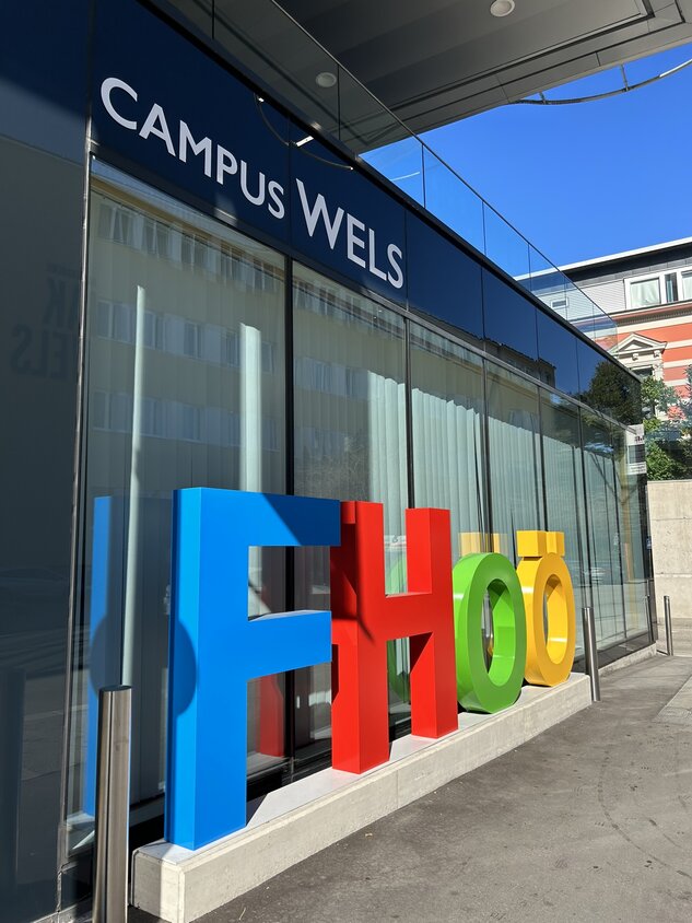 Der Eingang der Fachhochschule Wels. Vor der Glasfassade steht in bunden 3-D-Buchstaben der Schriftzug "FH OÖ", ober halt steht auf blauem Hintergrund in weißer Schrift "Campus Wels".